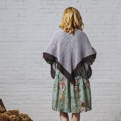 a woman standing in front of a white brick wall wearing a floral dress and shawl