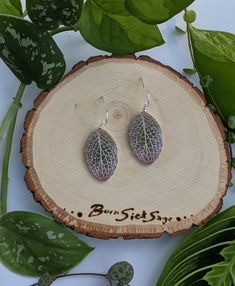 two leaf shaped earrings sitting on top of a piece of wood next to green leaves