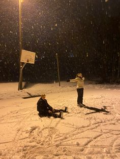 two people sitting in the snow at night with one person holding up a basketball hoop