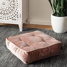 a square dog bed on the floor in front of a potted plant and rug