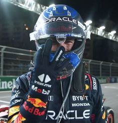 a man wearing a helmet and gloves talking on his cell phone while standing in front of a race car