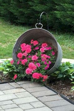 some pink flowers are in a bucket on the side of a brick walkway near grass and trees