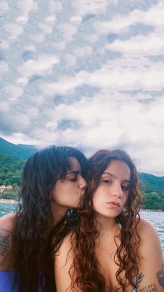 two beautiful young women standing next to each other near the water with mountains in the background