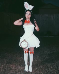 a woman dressed in bunny ears and white dress holding a clock with her right hand