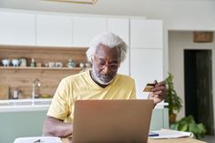 an older man sitting at a table with a laptop and credit card in front of him