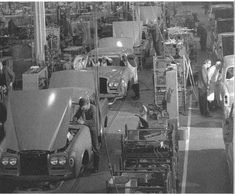 an assembly line with cars being worked on in the background and men standing around looking at them