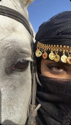 a woman wearing a black head covering next to a white horse with gold jewelry on it's forehead