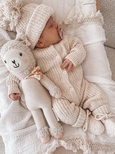 a baby laying on top of a bed next to a teddy bear wearing a knitted hat