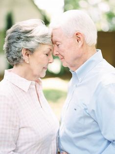 an older man and woman standing next to each other in front of a tree with their eyes closed