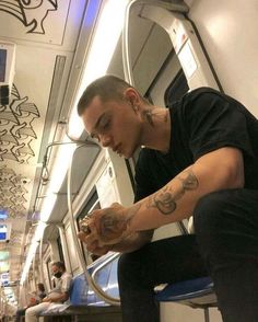 a man with tattoos sitting on a train looking at his cell phone while holding something in his hand