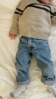 a baby laying on top of a white bed next to a stuffed teddy bear wearing a sweater and jeans