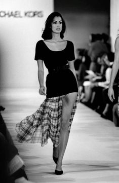 a woman is walking down the runway wearing a skirt and black shirt with plaid print on it