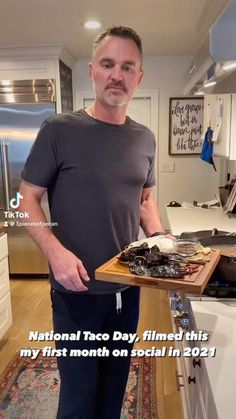 a man standing in a kitchen holding a tray with food on it that says national taco day, filmed this my first month on social in 2012