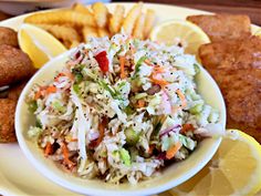 a white bowl filled with cole slaw next to some fried fish and lemon wedges