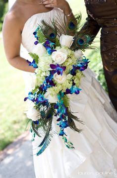 the bride is holding her blue and white wedding bouquet with peacock feathers on it's side