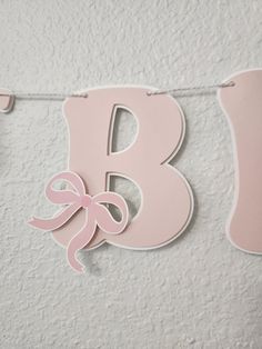 the letter b is decorated with pink ribbon and bows on a white wall hanging from a string