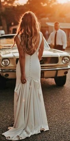 a woman in a white dress standing next to a car with her back turned towards the camera