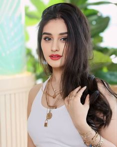 a woman with long dark hair wearing a white tank top and gold necklaces on her neck