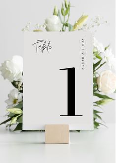 a white table number with flowers in the background and a wooden block for place card holders