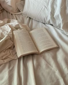 an open book laying on top of a bed with white sheets and pillows in front of it