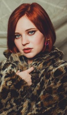 a woman with red hair and blue eyes wearing a leopard print fur coat is looking at the camera