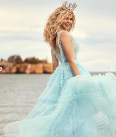 a woman in a blue dress is standing by the water with a tiara on her head