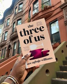 a person holding up a book about the three of us in front of some buildings