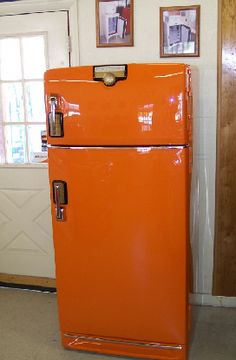 an orange refrigerator sitting in the corner of a room with pictures on the wall behind it