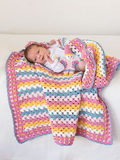 a baby laying on top of a blanket next to a white bed covered in a pink, yellow and blue crocheted blanket