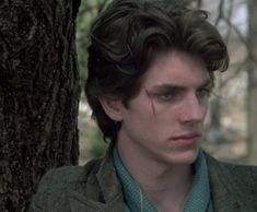 a young man standing next to a tree wearing a green sweater and blue collared shirt