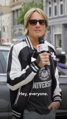 a man with sunglasses holding a microphone in front of a car and talking to someone on the street