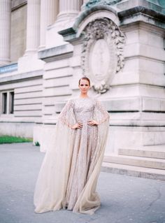 a woman standing in front of a building wearing a long dress with a cape on it