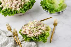 two bowls filled with lettuce and tuna salad next to gold forks on a marble surface