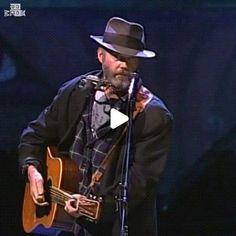 a man wearing a hat and holding a guitar in front of a microphone on stage