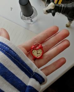 a person's hand with a small red heart shaped clock ring in front of a cat