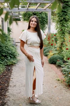 a woman wearing a white dress and sandals standing in front of some plants with her hands on her hips