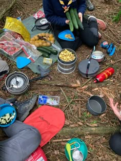 two people sitting on the ground with food in their hands and camping gear around them