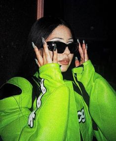 a woman in green jacket and sunglasses holding her hands to her face with one hand