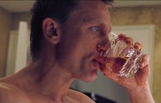 a man drinking water from a glass in the bathroom with only his shirtless body visible