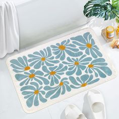 a bath mat with blue flowers on it next to slippers and a potted plant