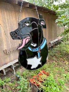 a stained glass dog hanging from a chain in a yard with flowers and trees behind it