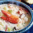 a bowl of lobster chowee on a blue tablecloth with bread in the background