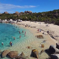 many people are on the beach and in the water near some large rocks, with clear blue water