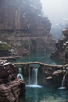 a small bridge over a body of water in front of a rocky cliff face with a waterfall