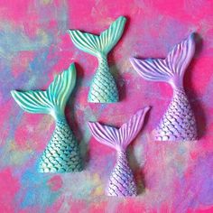 three little mermaid tail ornaments sitting on top of a pink surface with blue and green paint