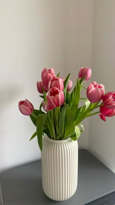 a white vase filled with pink tulips on top of a gray shelf next to a wall