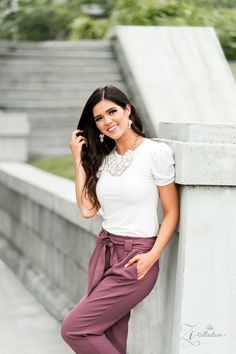 a beautiful woman leaning against a wall wearing purple pants and a white top with pearls on her neck