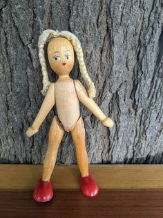 a wooden doll with blonde hair and red shoes standing in front of a tree trunk