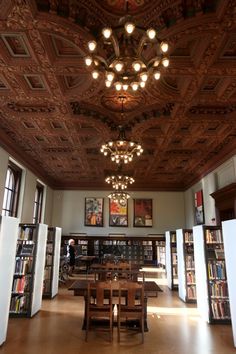 an ornately decorated library with tables and bookshelves in the center is lit by chandeliers