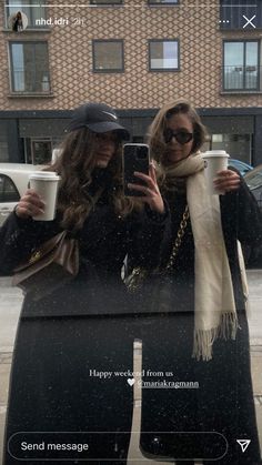 two women standing in front of a car holding coffee cups and looking at their cell phone